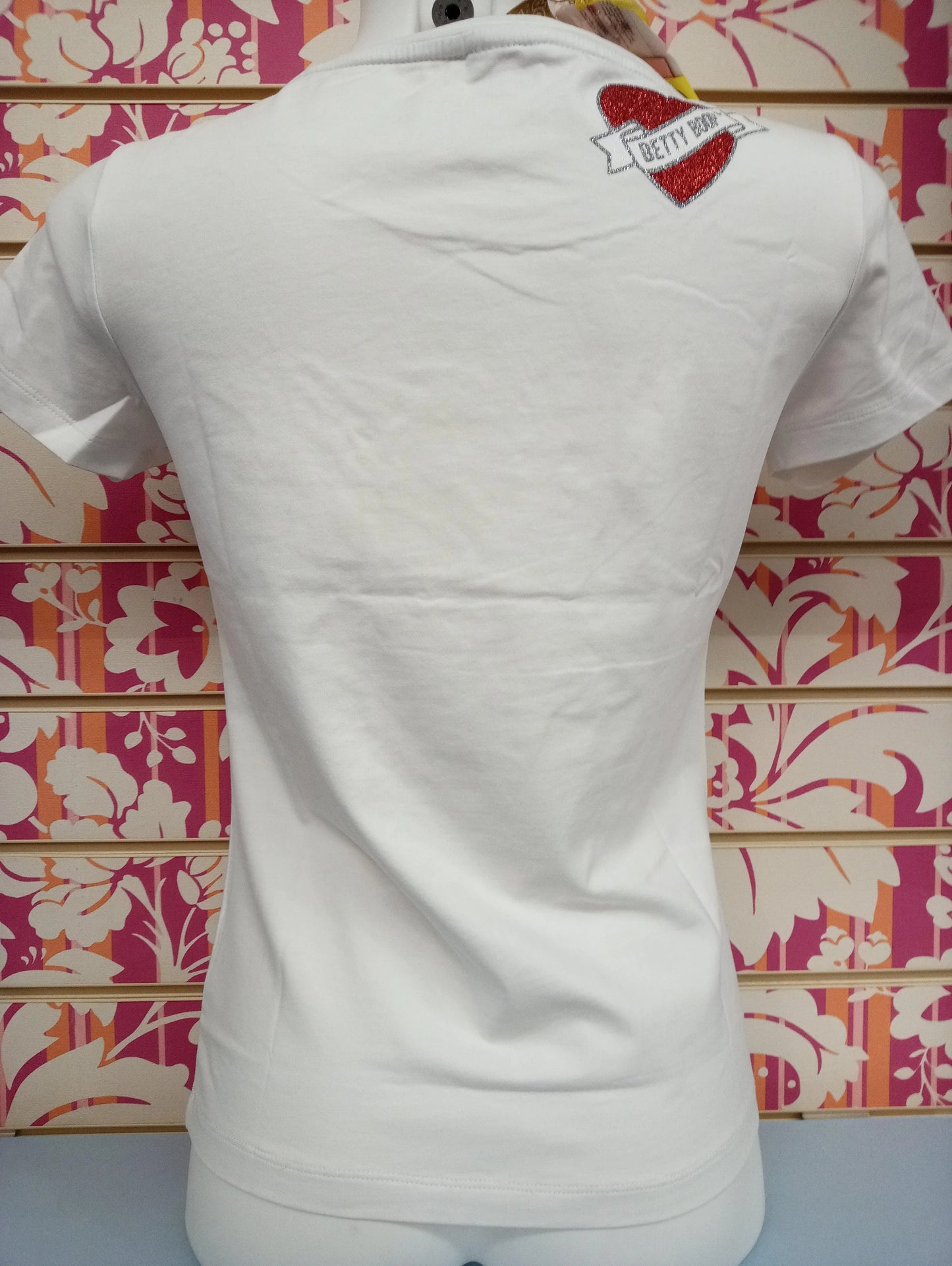 T-shirt donna bianca in cotone, stile streetwear, dettaglio cuore glitterato rosso con scritta Betty Boop sulla spalla, fondo neutro a motivi floreali.
