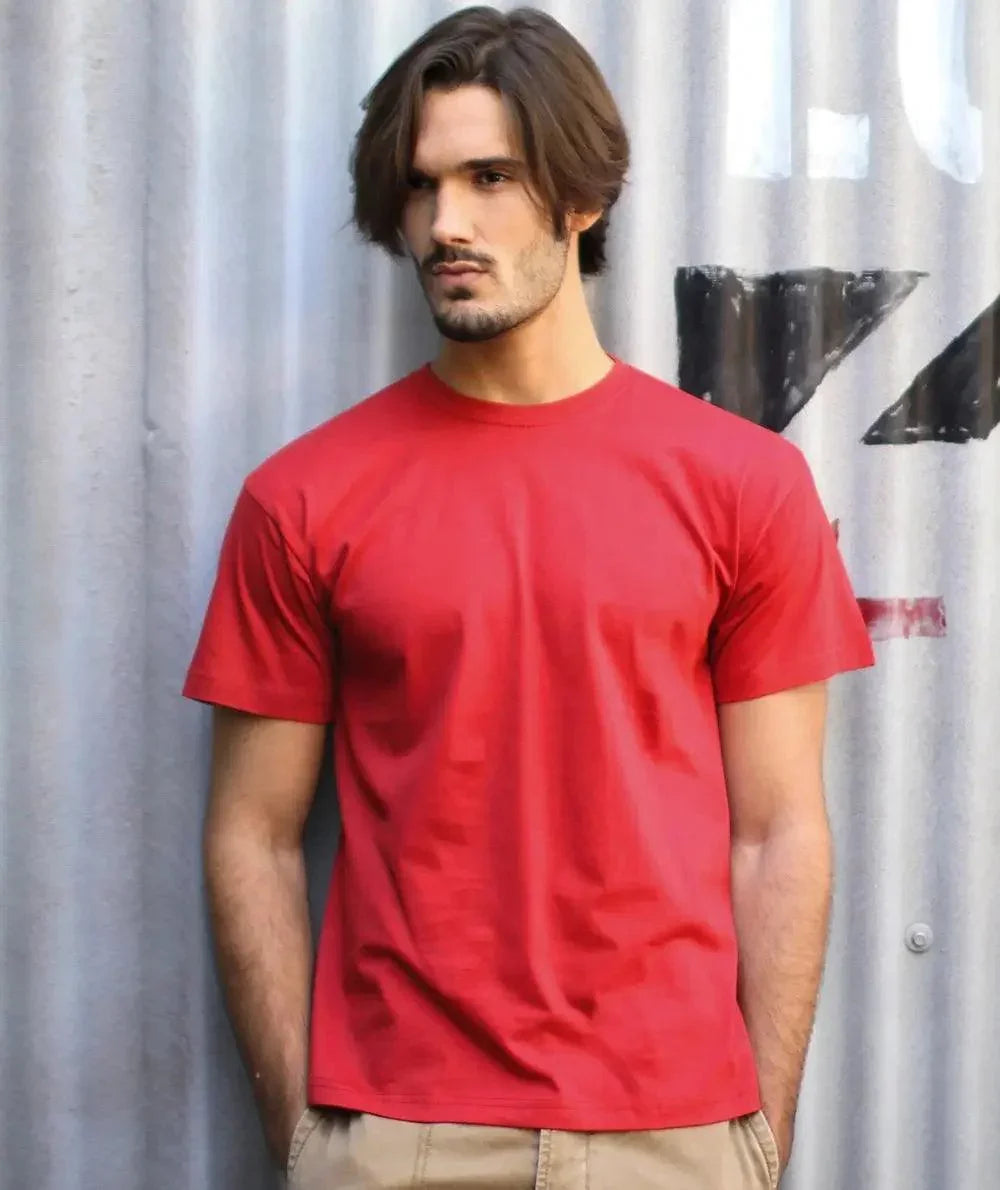 Uomo con t-shirt rossa in cotone, stile streetwear urban, davanti a parete industriale.
