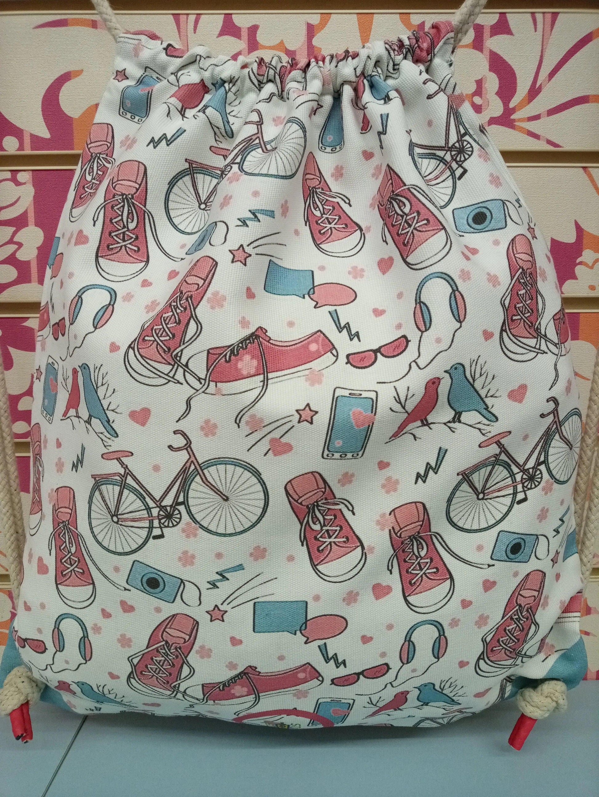 Zaino a sacca streetwear in tessuto bianco con stampe urban colorate di scarpe, bici, cuffie e dettagli rosa e azzurri.
